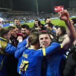 A group of soccer players in blue and yellow celebrate together on the field, fists raised, with cheering fans in the stands behind them.