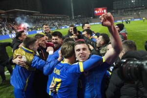 A group of soccer players in blue and yellow celebrate together on the field, fists raised, with cheering fans in the stands behind them.