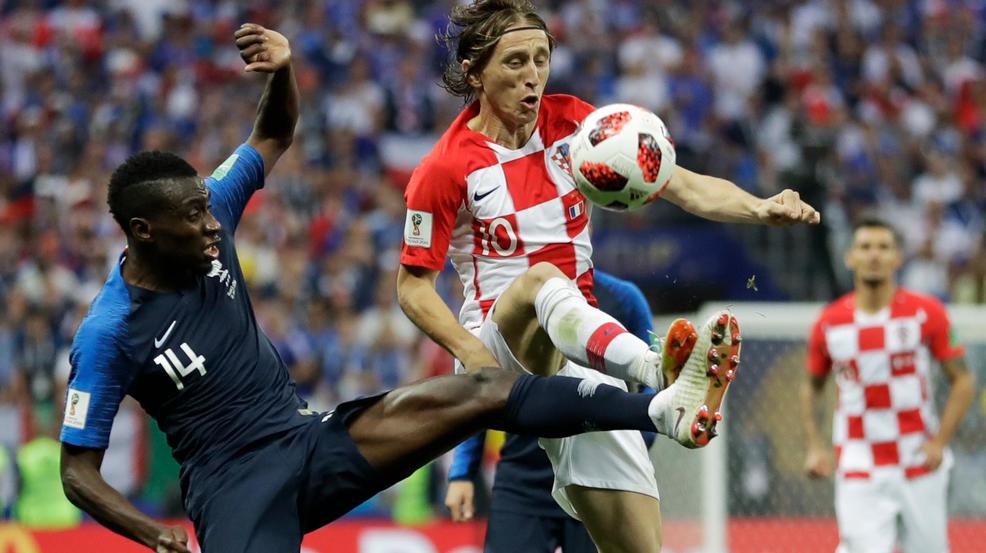 Croatian player in red‑and‑white checkered kit and French defender in blue compete for the ball in midair during a soccer match.