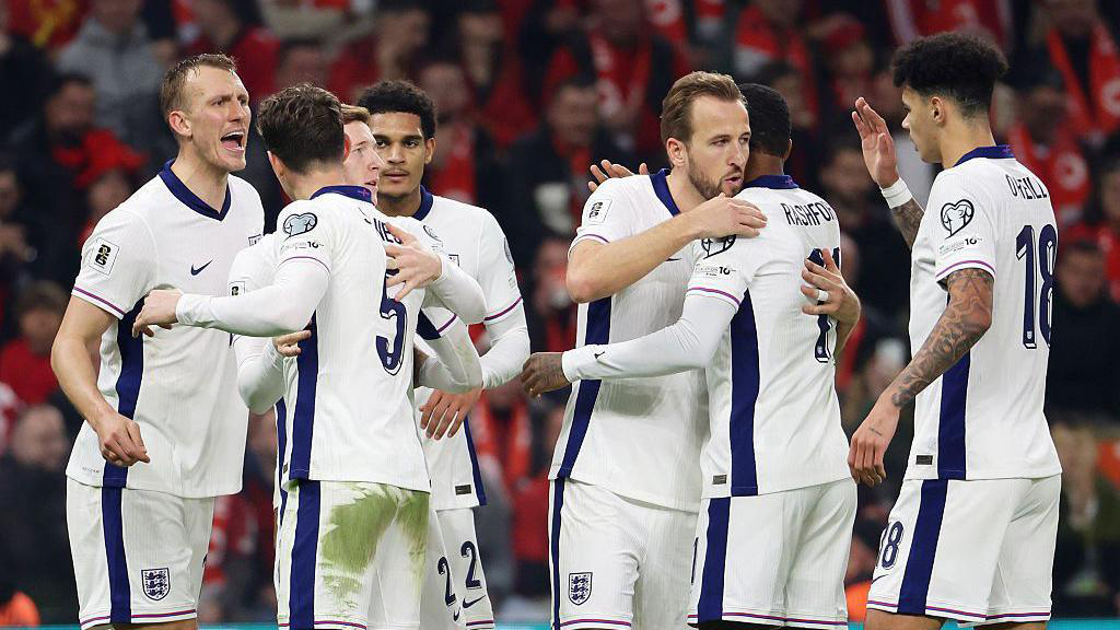England football players in white kits celebrate together on the pitch after a goal with fans in a red stadium behind them.