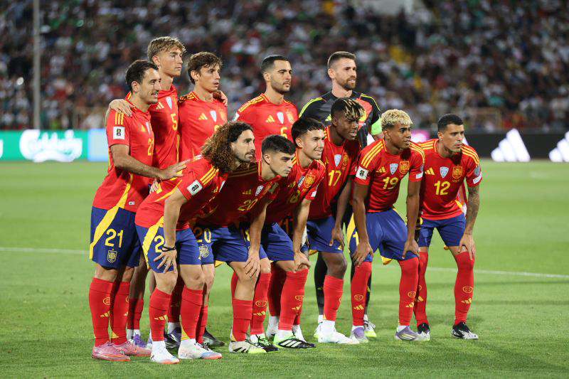 Spain men's national football team players posing for a lineup photo on the pitch before a match in red jerseys and blue shorts.