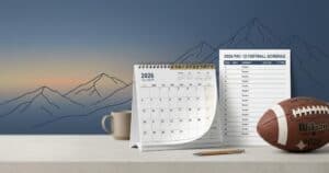 Desk with a 2026 calendar, football schedule, Wilson football, a pencil, and a mug against a mountain backdrop.