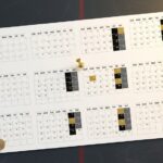 Top-down view of a desk calendar sheet showing twelve monthly grids arranged in a 3x4 layout, with a yellow pushpin marking one month; a wooden spoon and a black round cap sit on a dark surface nearby.